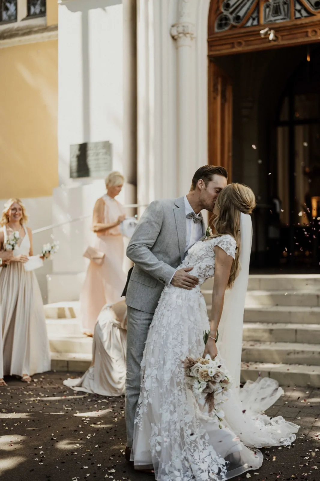 Philina & Damian – Ein frisch verheiratetes Paar küsst sich vor einer Kirche, während im Hintergrund Brautjungfern stehen
