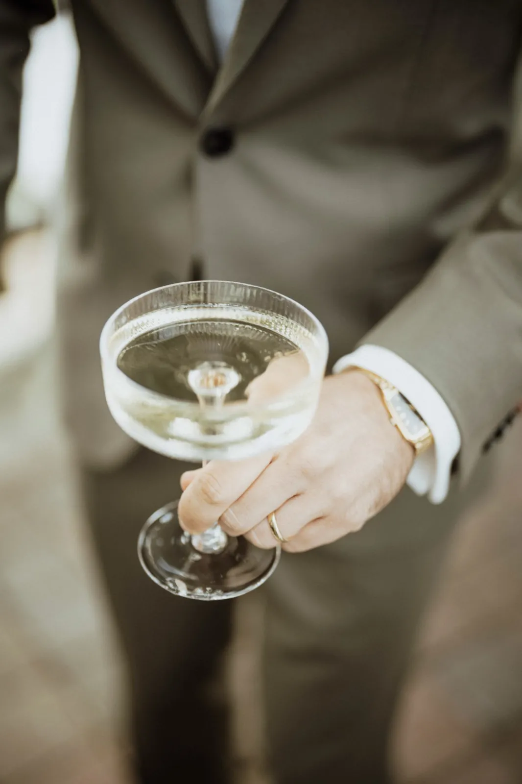Luisa & Norman – Ein Mann im Anzug hält ein Champagnerglas, an seiner Hand ist ein Ehering sichtbar