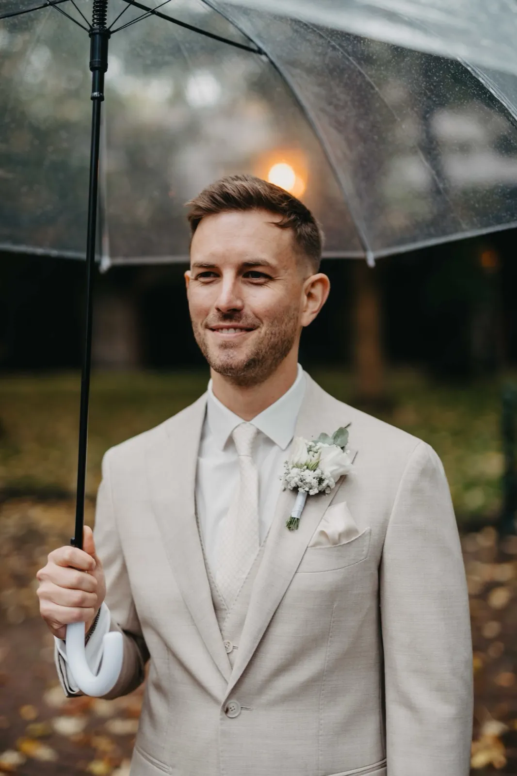 Alina & Timo – Ein Bräutigam im beigen Anzug lächelt unter einem transparenten Regenschirm im Freien
