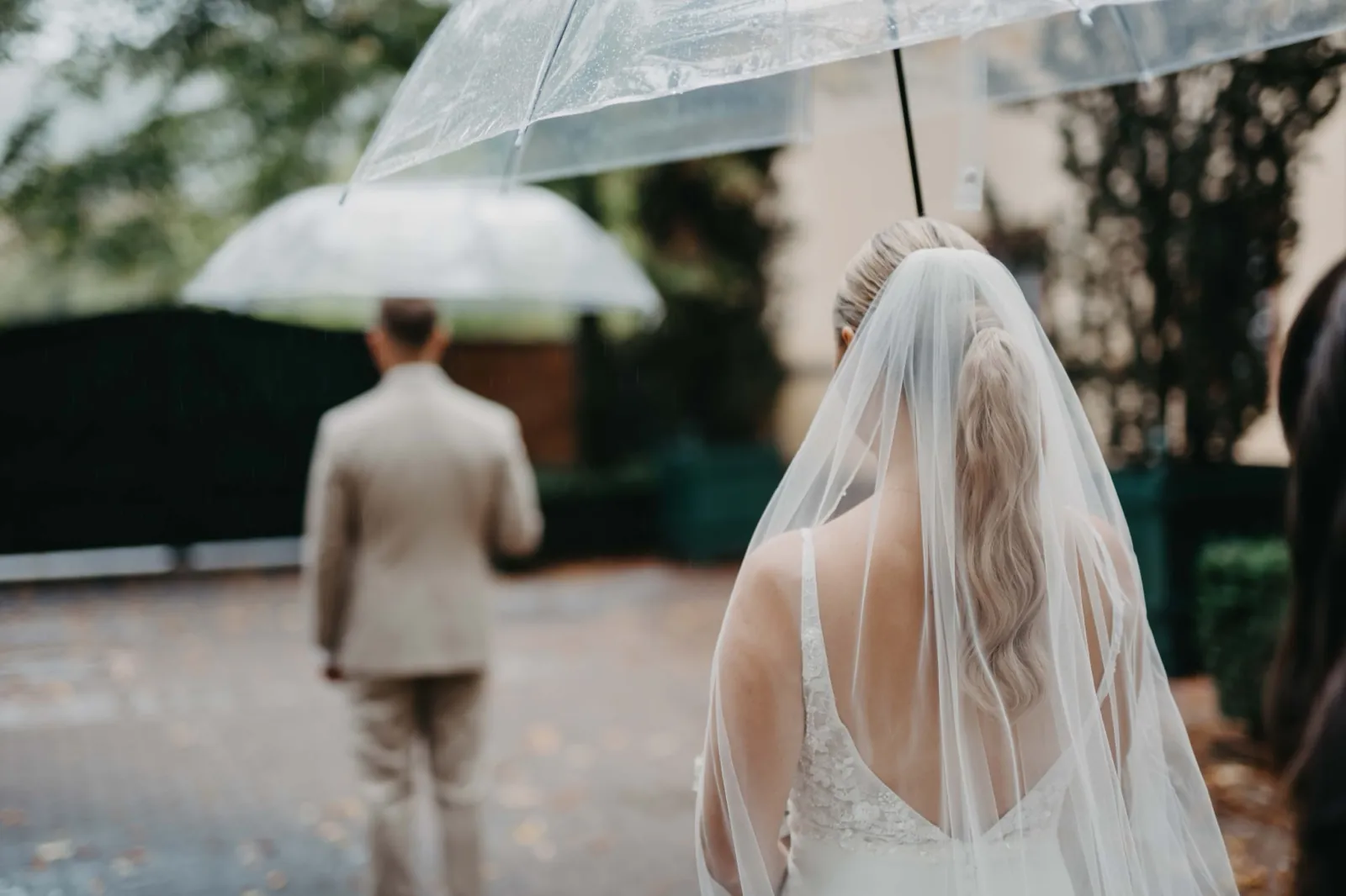 Alina & Timo – Eine Braut nähert sich von hinten dem Bräutigam im Regen unter Regenschirmen