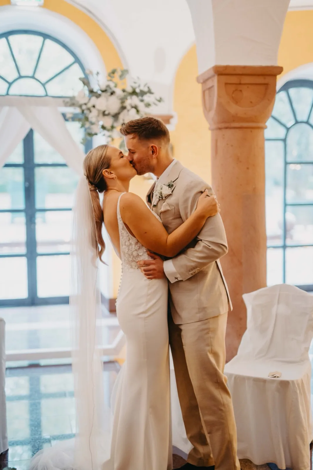 Alina & Timo – Ein Brautpaar küsst sich innig während der Hochzeitszeremonie in einem hellen Innenraum