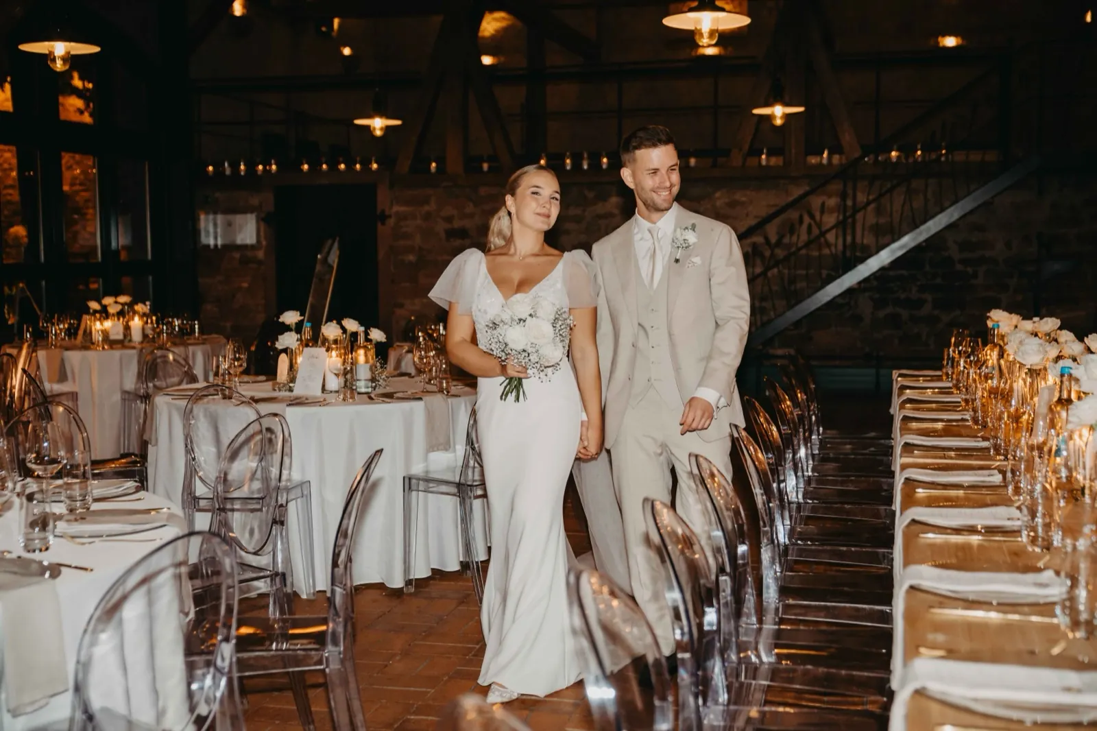 Alina & Timo – Ein Brautpaar läuft lächelnd durch einen festlich geschmückten Hochzeitssaal mit gedeckten Tischen
