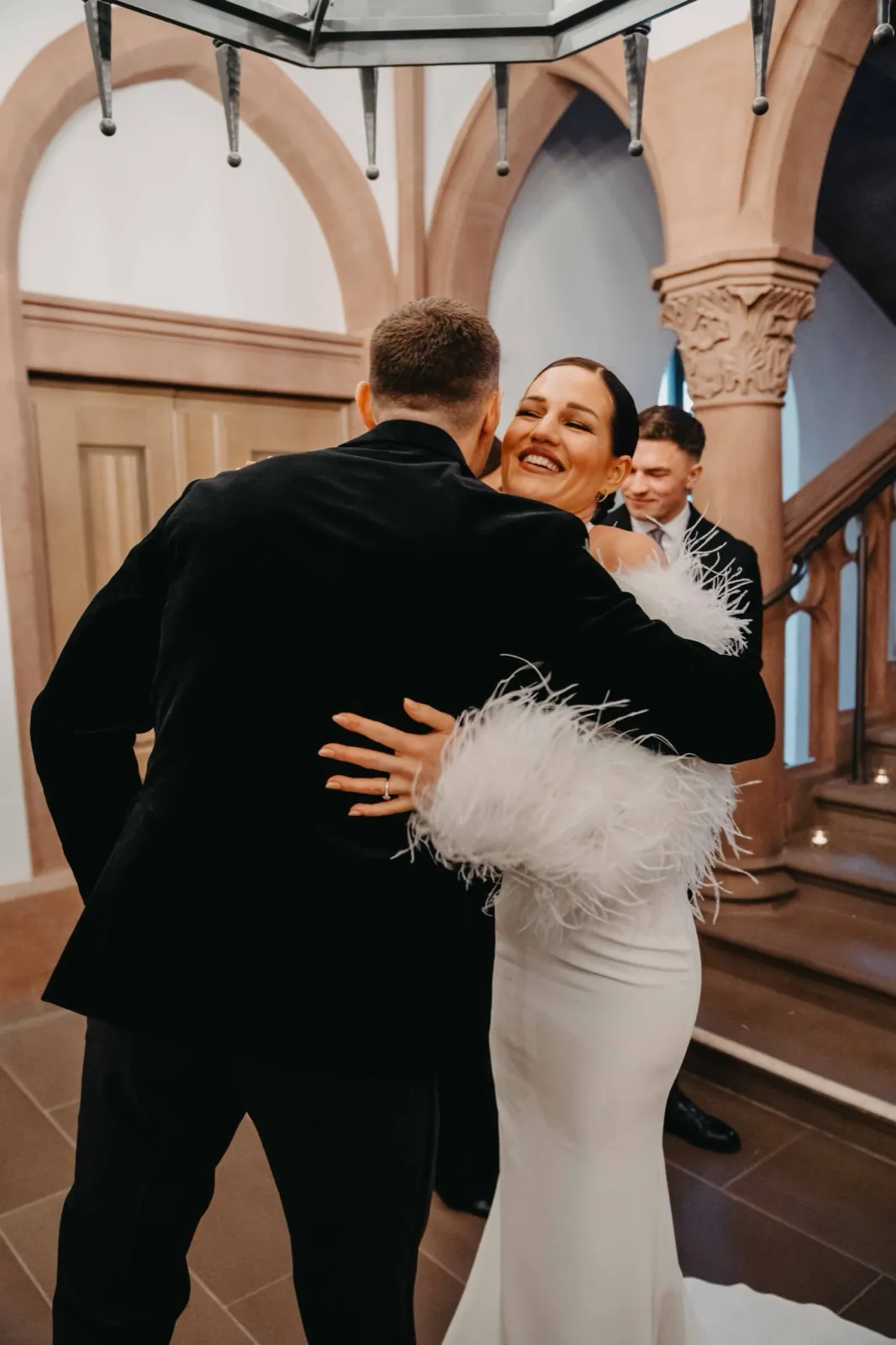 Alisa & Lars – Eine Braut im weißen Kleid mit Federn umarmt den Bräutigam in einem historischen Gebäude
