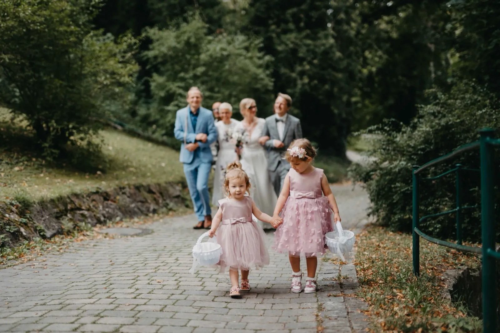 Barbara & Gabi – Zwei Blumenmädchen in rosa Kleidern laufen vor der Hochzeitsgesellschaft auf einem gepflasterten Weg