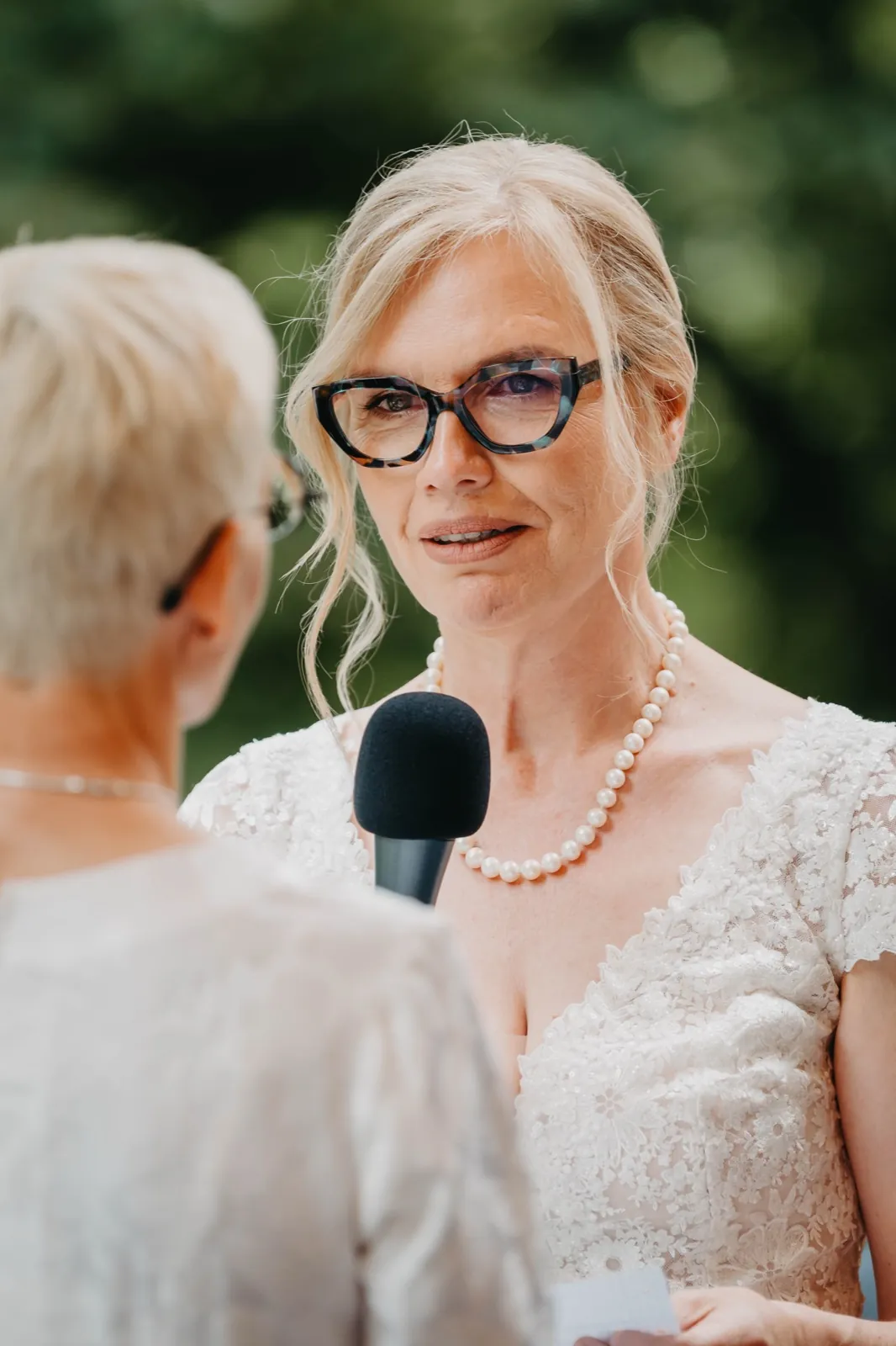 Barbara & Gabi – Eine Braut mit Brille und Perlenkette spricht während der Trauung in ein Mikrofon