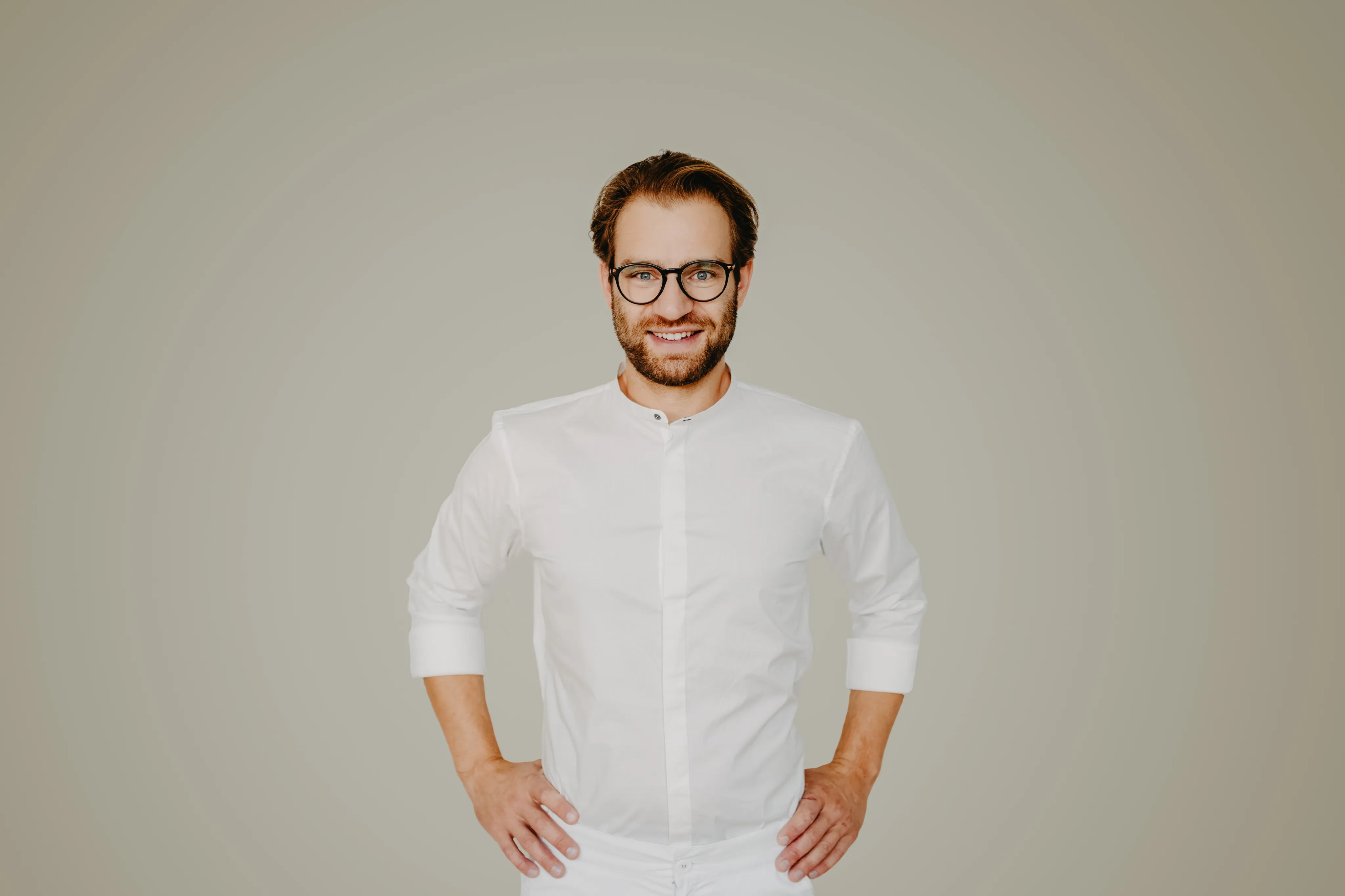 Bünger & Meißner – Ein lächelnder Mann mit Brille und weißem Hemd steht vor grauem Hintergrund