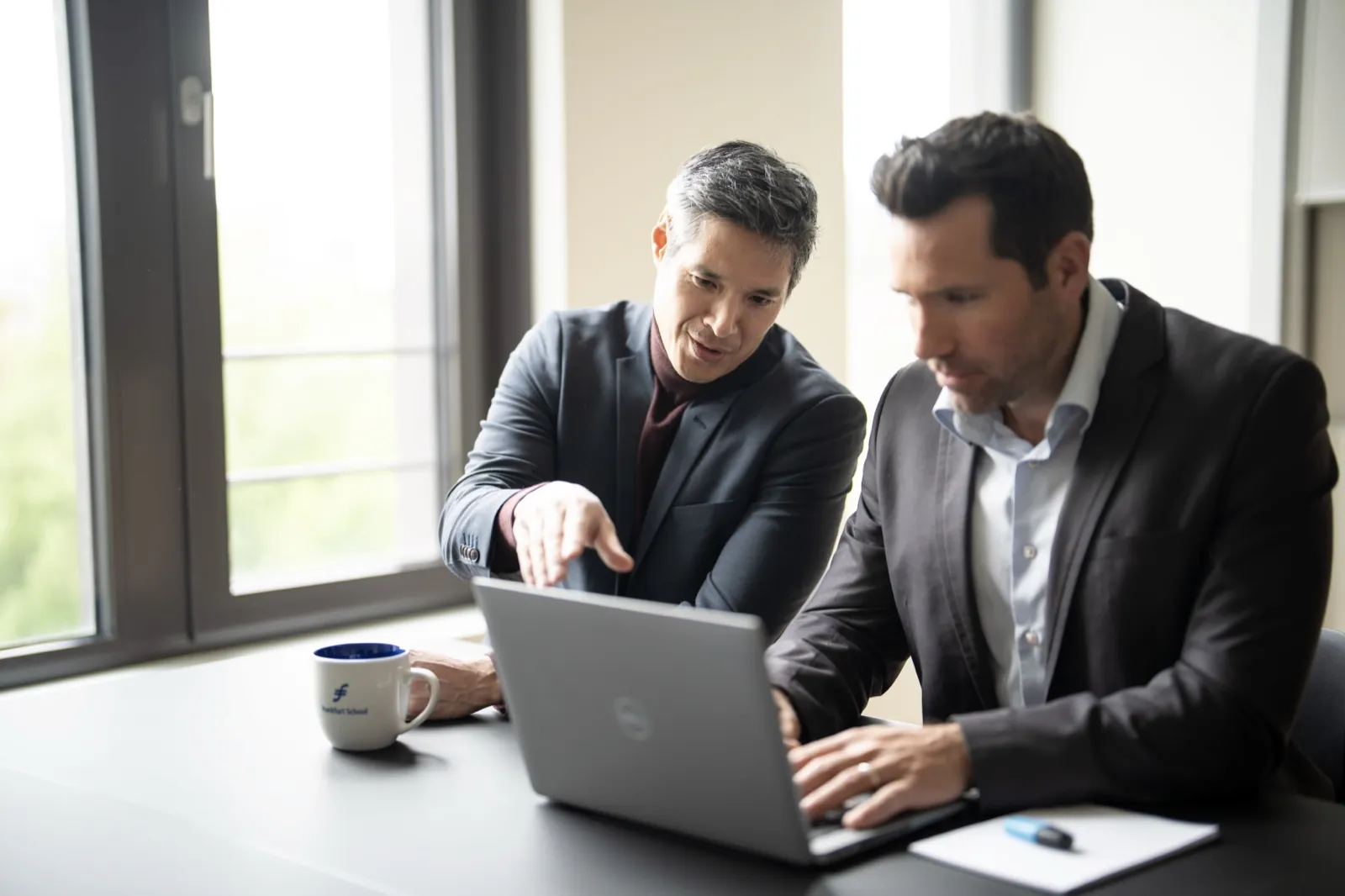 Frankfurt School – Zwei Männer im Anzug besprechen ein Dokument auf einem Laptop an einem Tisch