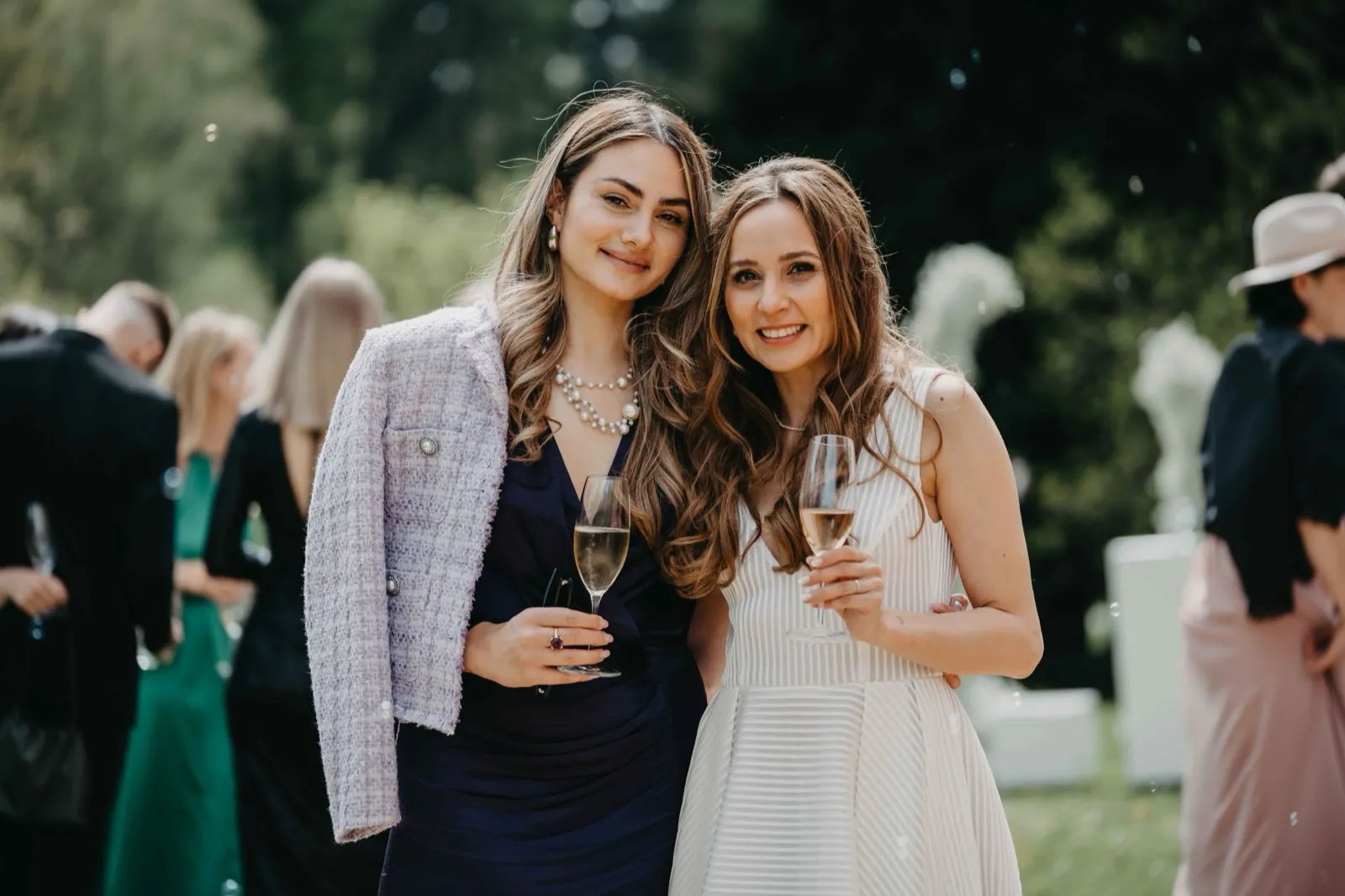 Imy & Felix – Zwei elegant gekleidete Frauen lächeln mit Champagnergläsern bei einer Feier im Freien