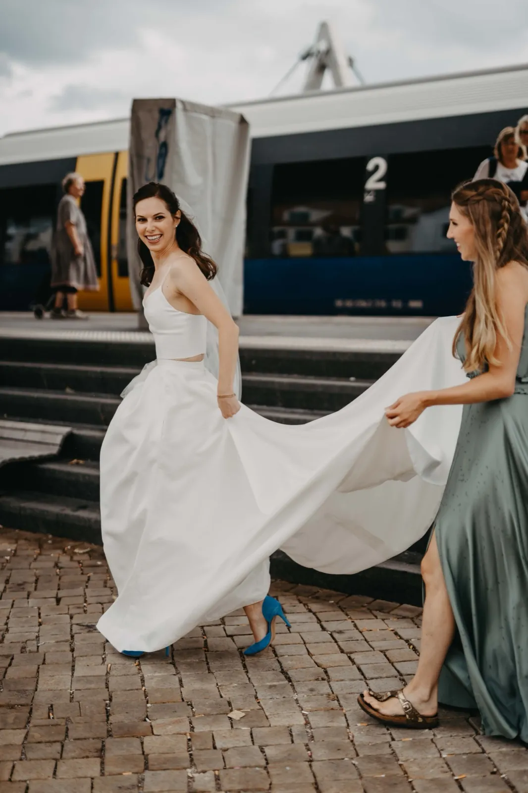 Isa & Nico – Eine Braut läuft lachend an einem Bahnhof, während eine Brautjungfer ihr Kleid hält