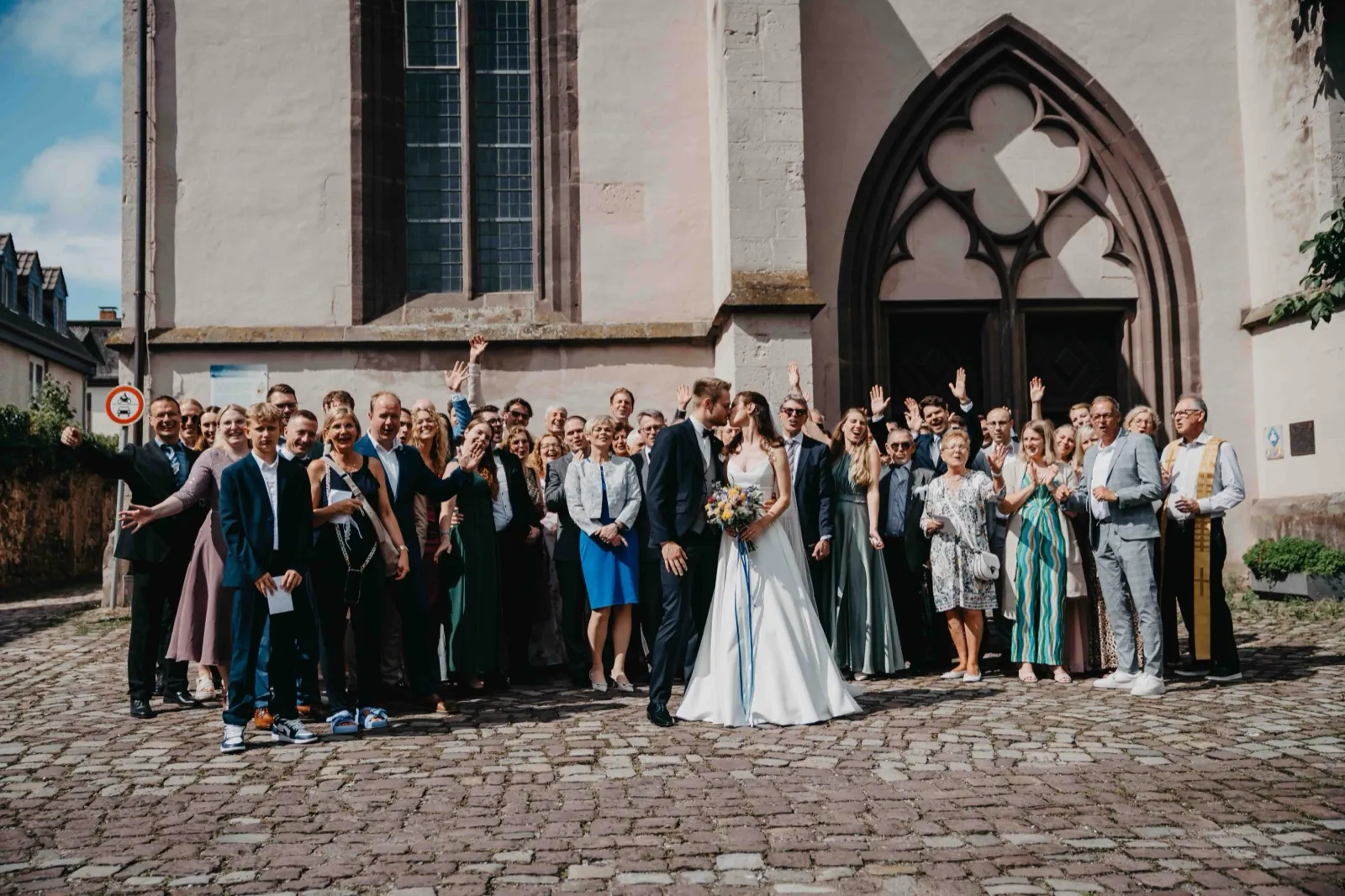 Isa & Nico – Eine große Hochzeitsgesellschaft posiert für ein Gruppenfoto vor dem Eingang einer Kirche