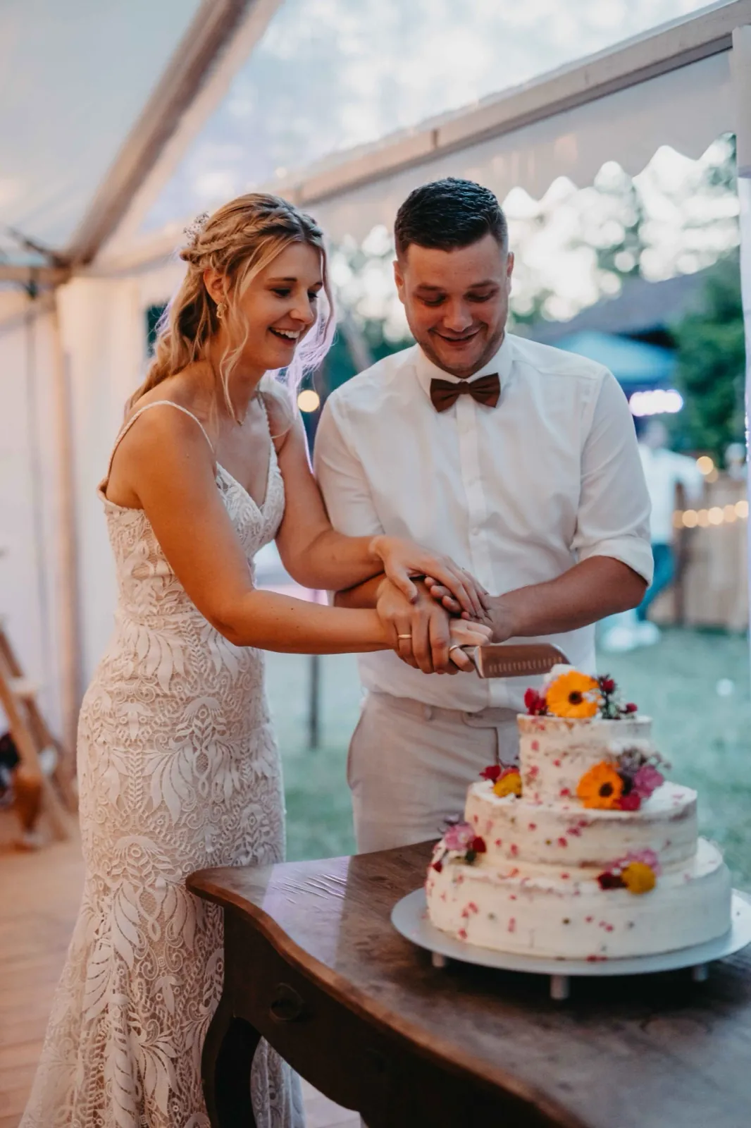 Jana & Christoph – Ein Brautpaar schneidet gemeinsam die dreistöckige, mit Blumen verzierte Hochzeitstorte an