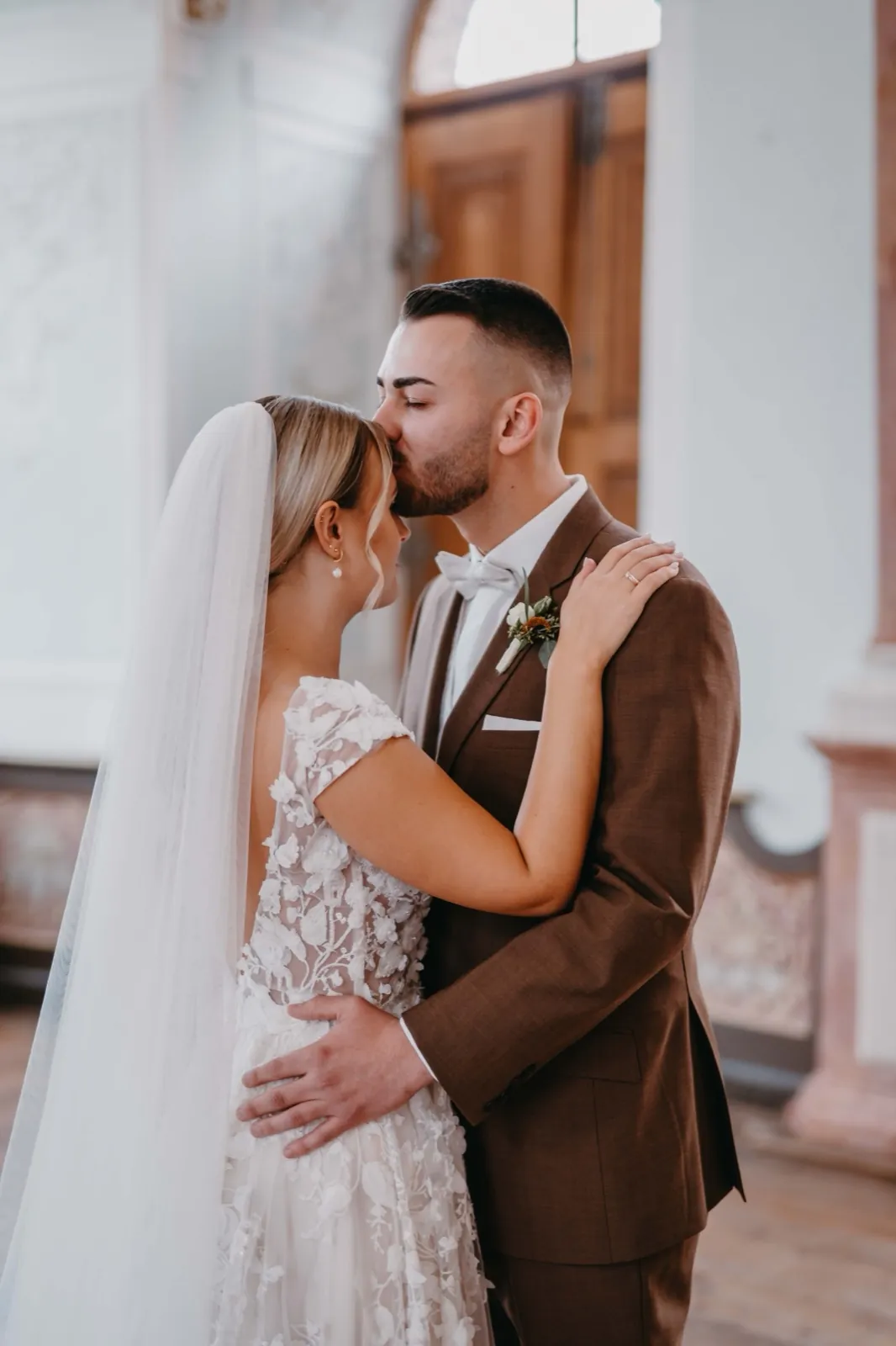 Jana & Daniel – Ein Bräutigam küsst seine Braut in einem hellen Innenraum auf die Stirn