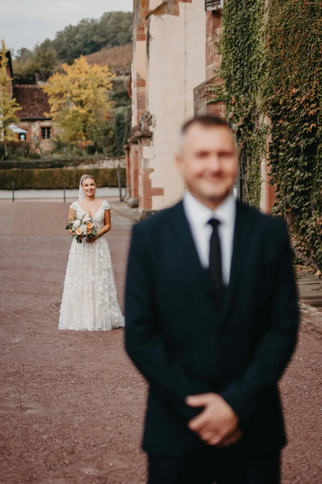 Jana & Daniel – Eine Braut steht mit einem Blumenstrauß im Hintergrund, während der Bräutigam im Vordergrund wartet