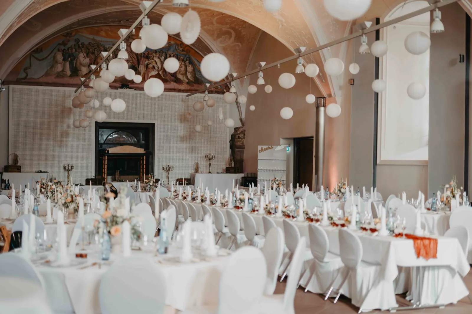 Jana & Daniel – Ein festlich dekorierter Hochzeitssaal mit weißen Tischen, Stühlen und hängenden Papierlaternen