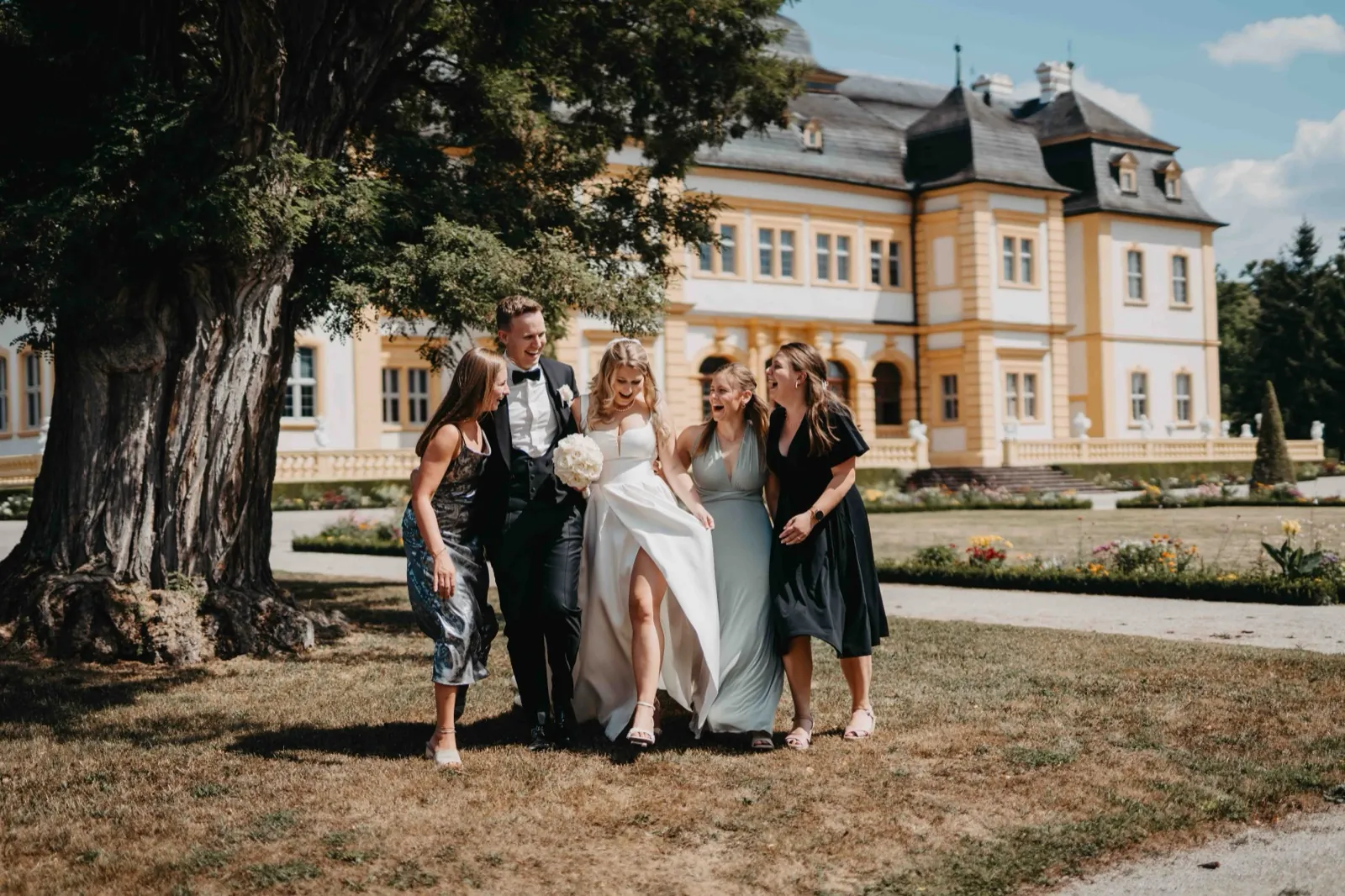 Janina & Alexander – Ein Brautpaar läuft lachend mit Gästen vor einem großen gelben Schloss im Freien