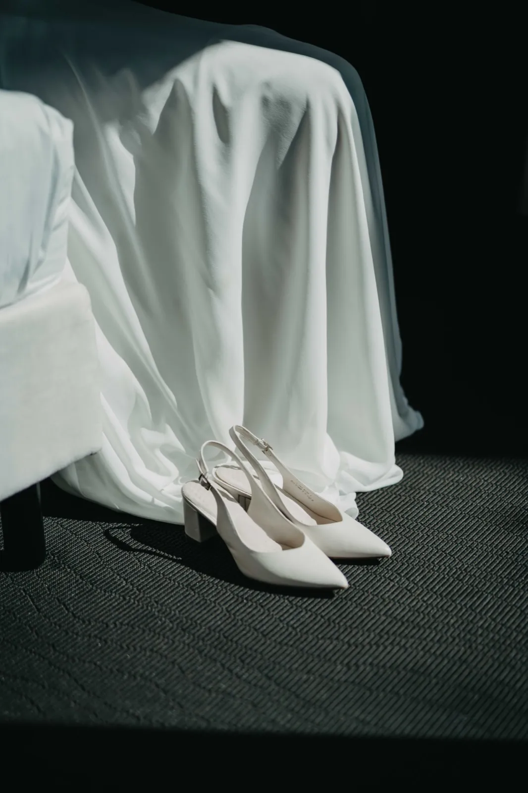 Julia & Max – Weiße Brautschuhe stehen auf einem dunklen Teppich vor einem weißen Brautkleid