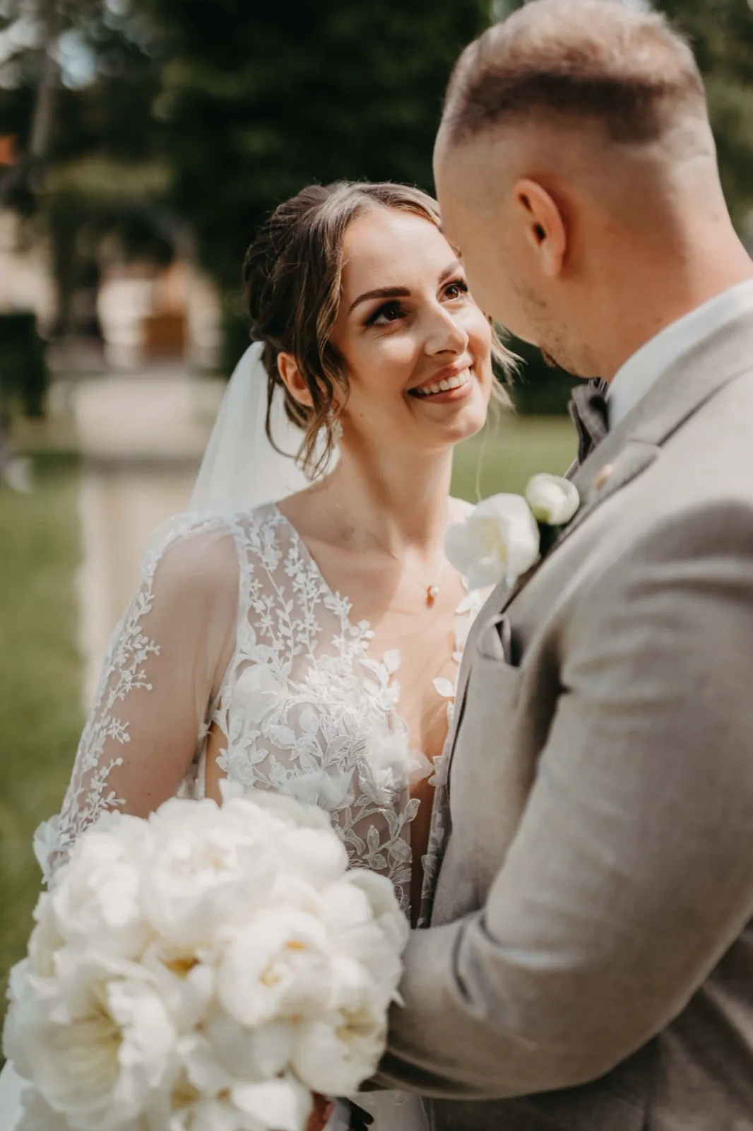 Kristina & Pascal – Ein Brautpaar blickt sich lächelnd an, während die Braut einen weißen Blumenstrauß hält