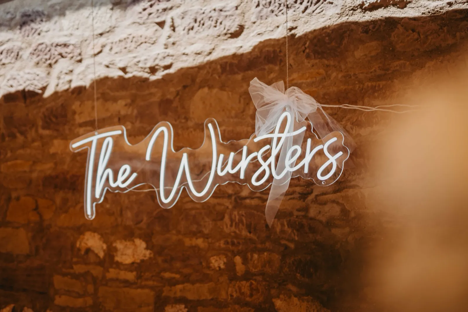 Kristina & Pascal – Ein leuchtendes Neonschild mit der Aufschrift „The Wursters“ hängt vor einer rustikalen Steinwand