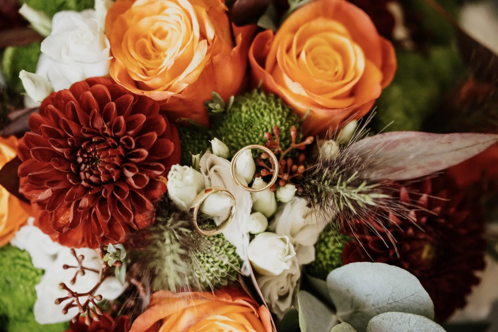 Marie & Thomas – Zwei goldene Eheringe liegen eingebettet in einem farbenfrohen Brautstrauß aus Rosen und Dahlien