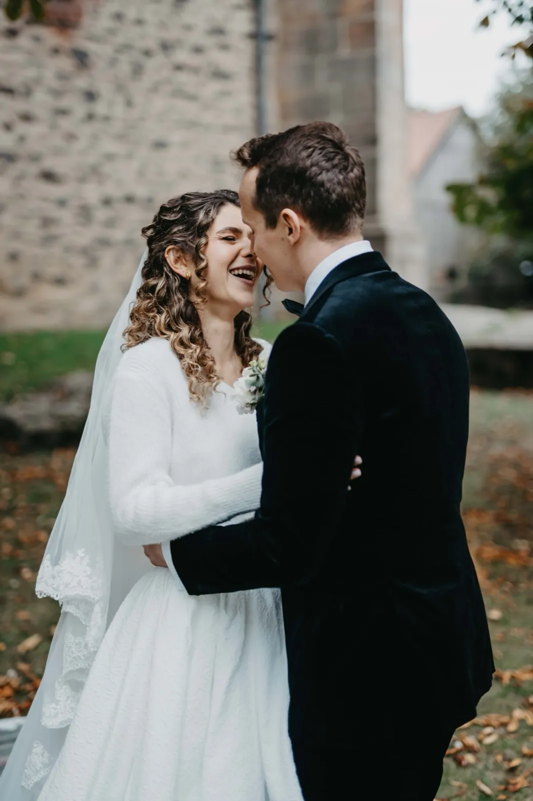Mia & Clemens – Ein lachendes Brautpaar umarmt sich im Freien vor einer steinernen Wand