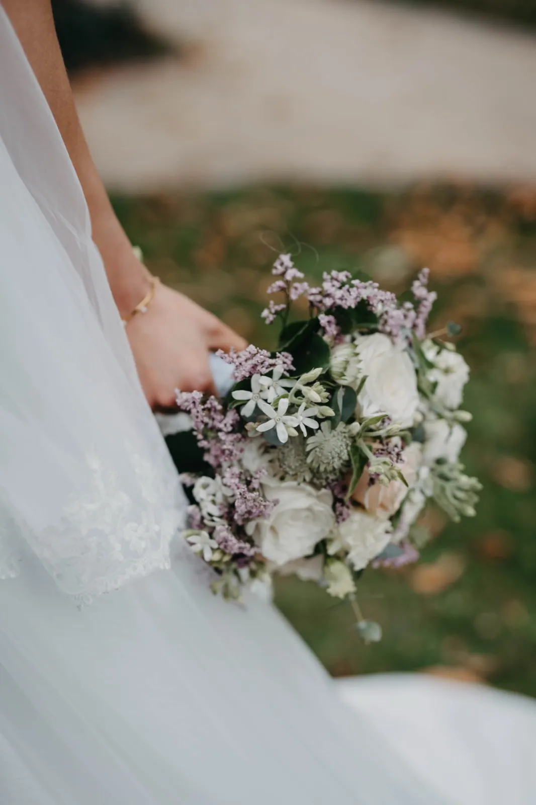 Mia & Clemens – Eine Braut hält einen Brautstrauß mit weißen Rosen und lila Blüten in der Hand