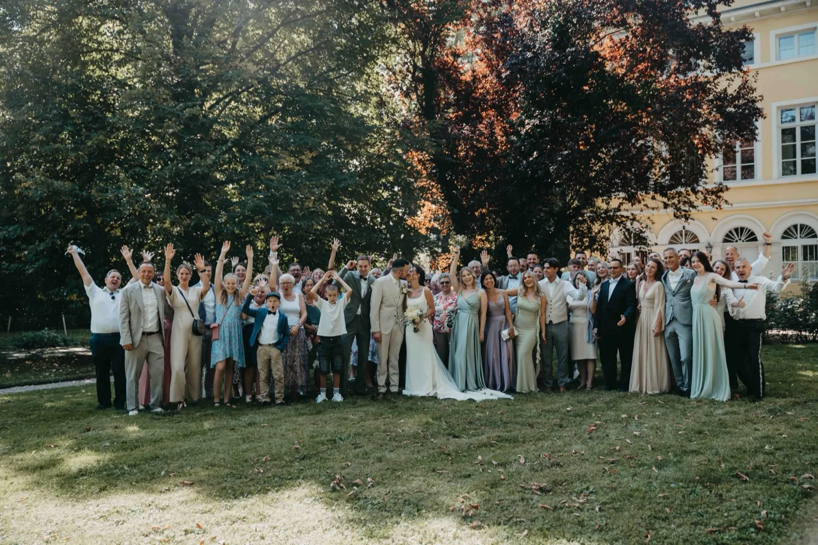 Michelle & Felix – Eine große Hochzeitsgesellschaft posiert für ein Gruppenfoto auf einer Wiese vor Bäumen