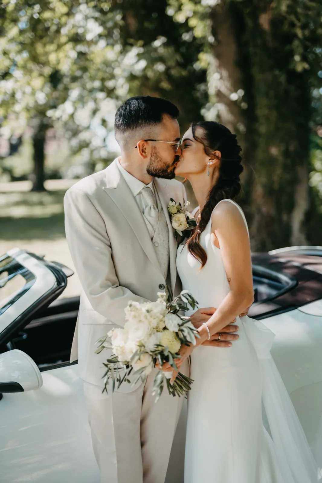 Michelle & Felix – Ein Brautpaar küsst sich im Freien neben einem weißen Cabriolet