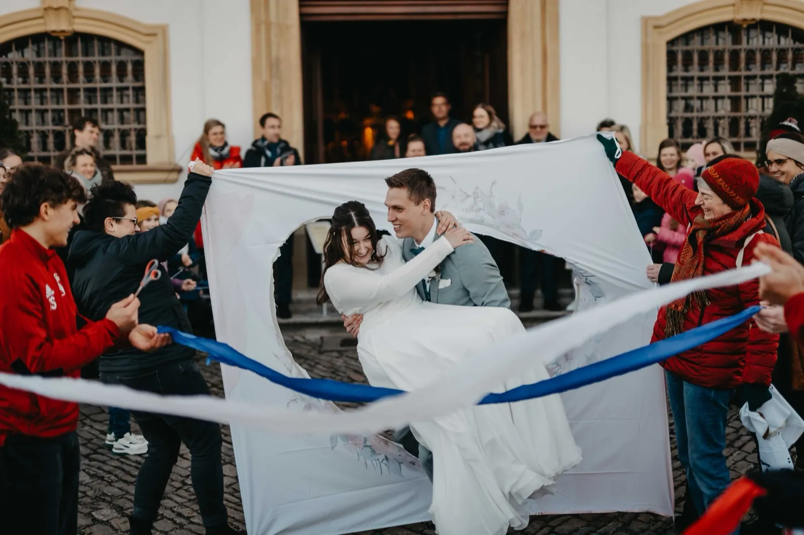Sophie & Mirko – Hochzeitsbrauch vor der Kirche: das Brautpaar am herzförmig ausgeschnittenen Laken, umringt von jubelnden Gästen